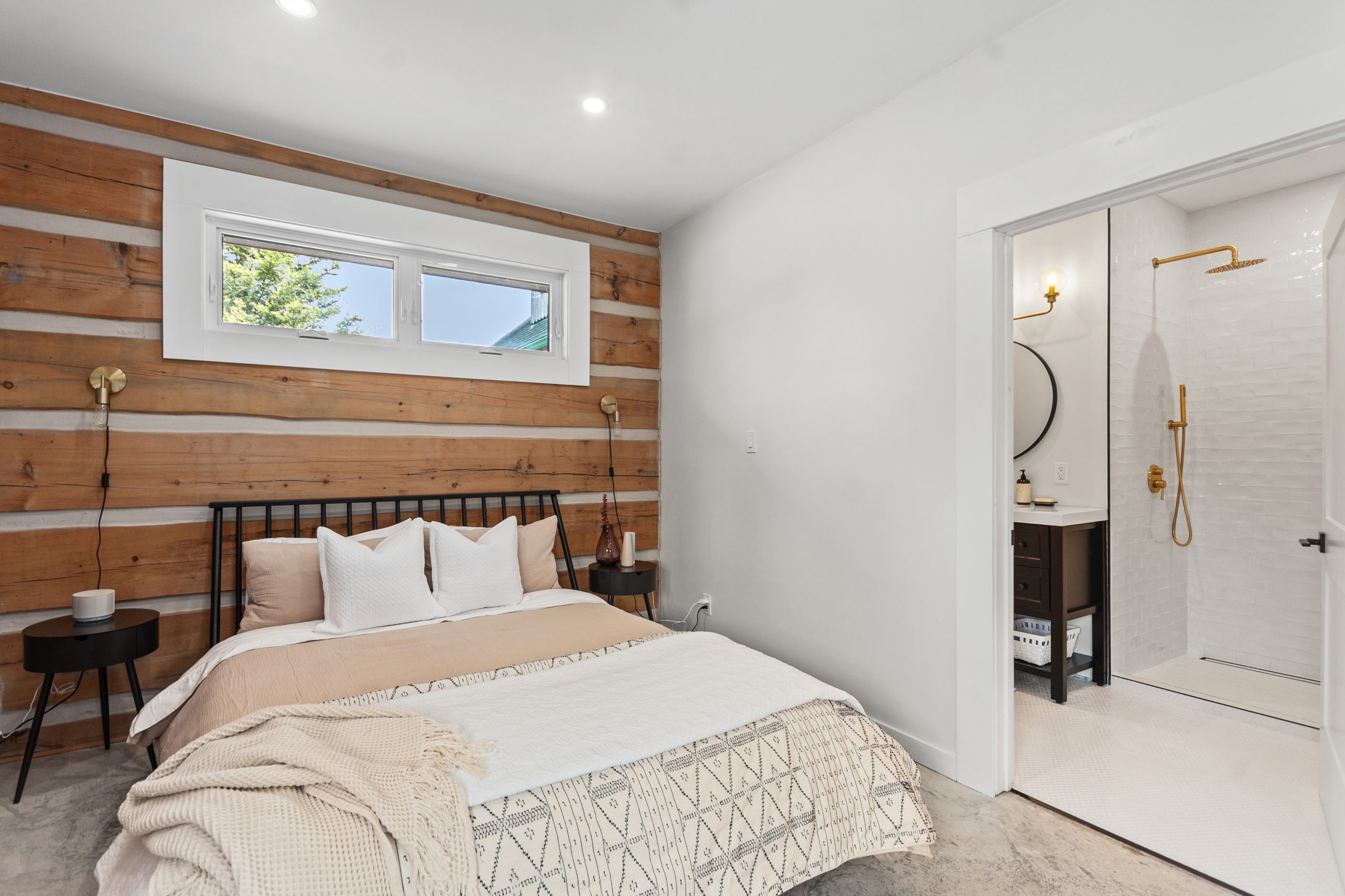 Bright bedroom with a double bed, log accent wall, and doorway leading to an ensuite bathroom.