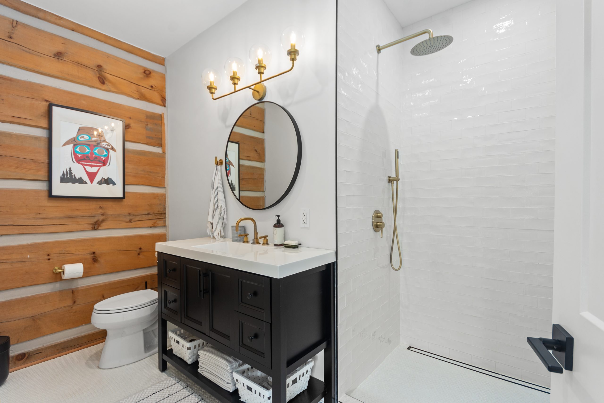 Bathroom with a walk-in shower, dark vanity with sink, round mirror, and log wall accent.
