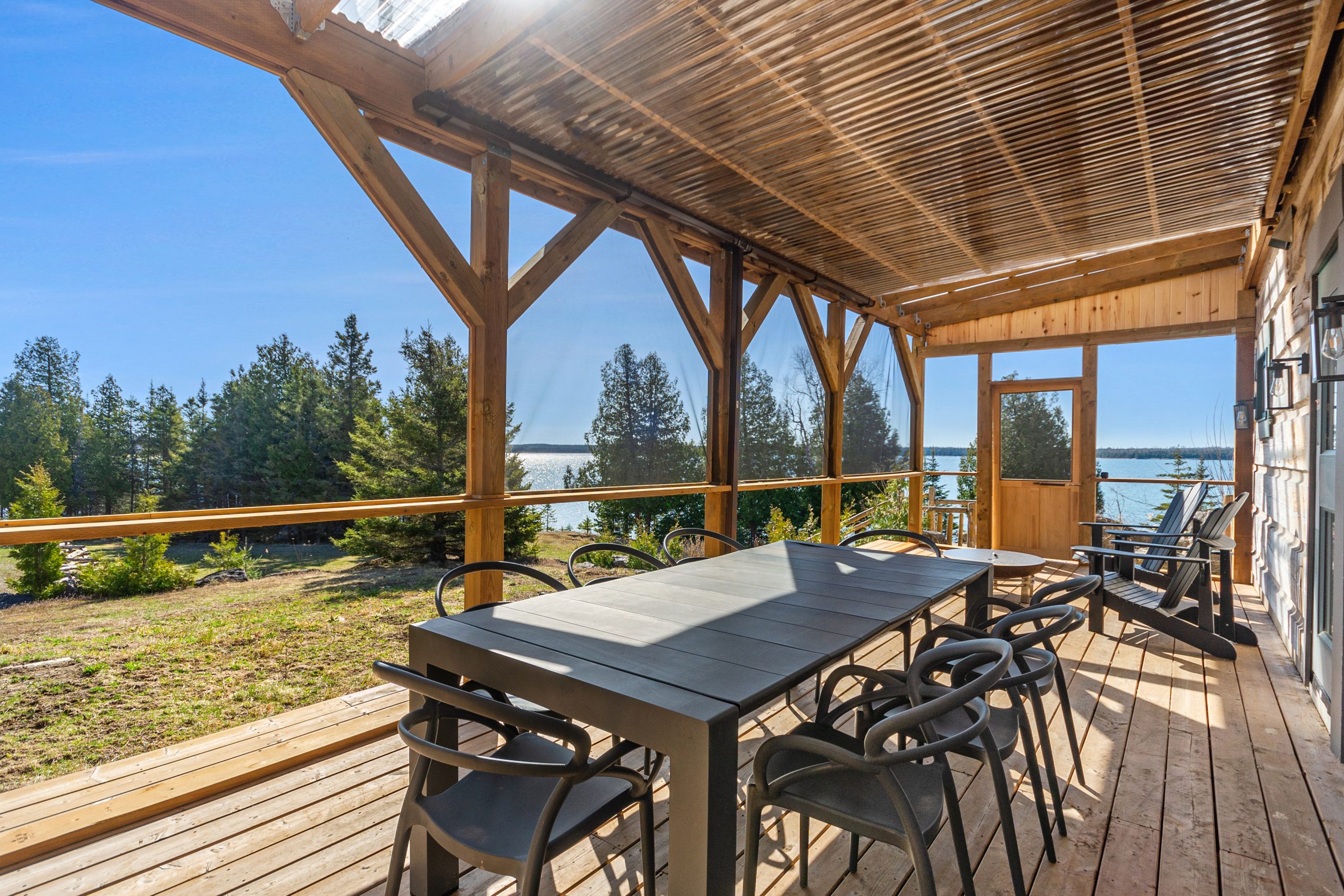 Covered deck with an outdoor dining table and chairs, looking out toward the water and treed shoreline.