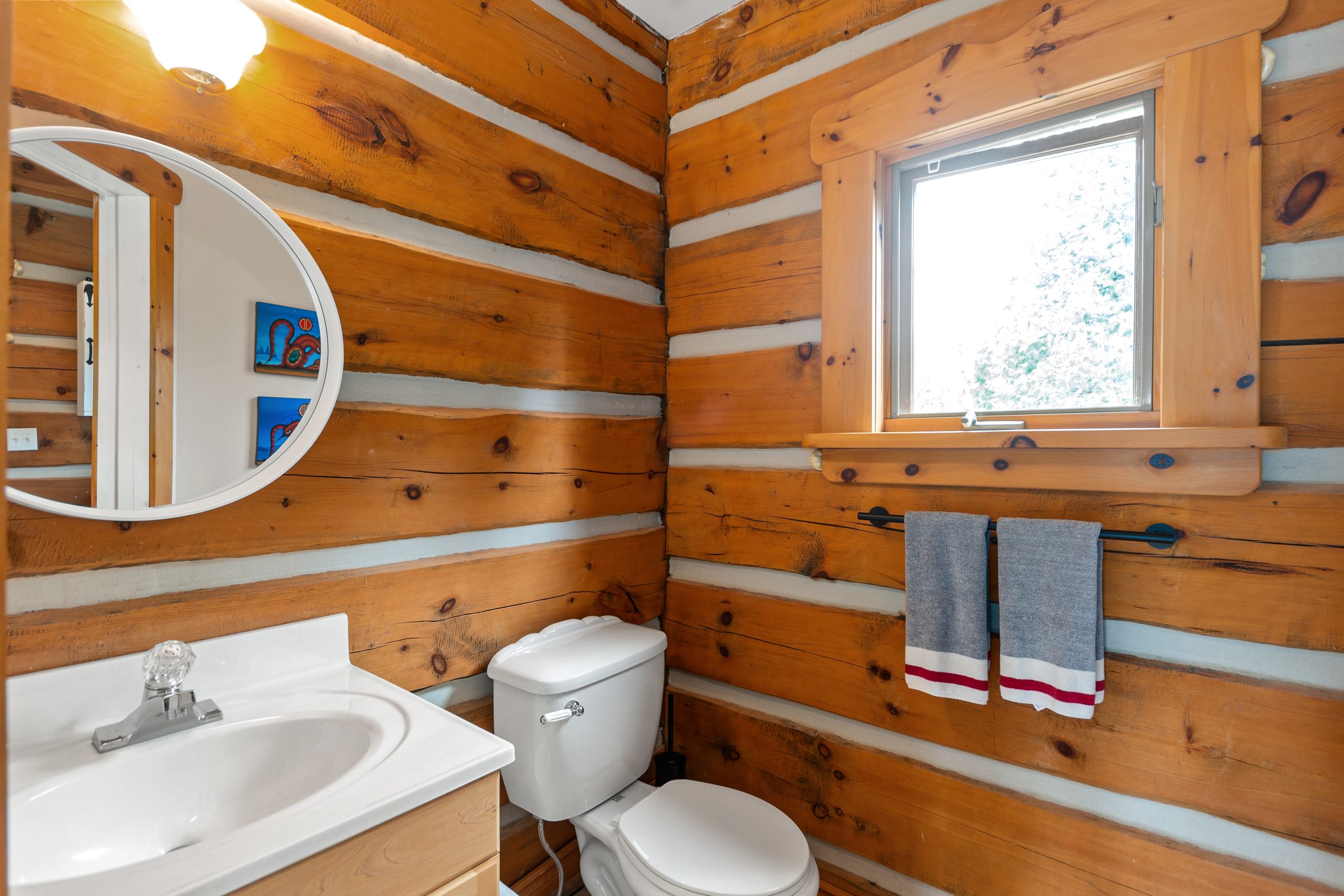 Bathroom with a vanity, toilet, and log walls, with a window above the sink.