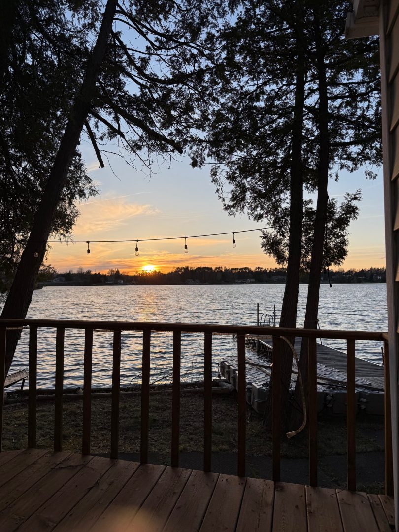 Sunset over Beaver Lake viewed from the cottage deck, framed by trees and string lights.