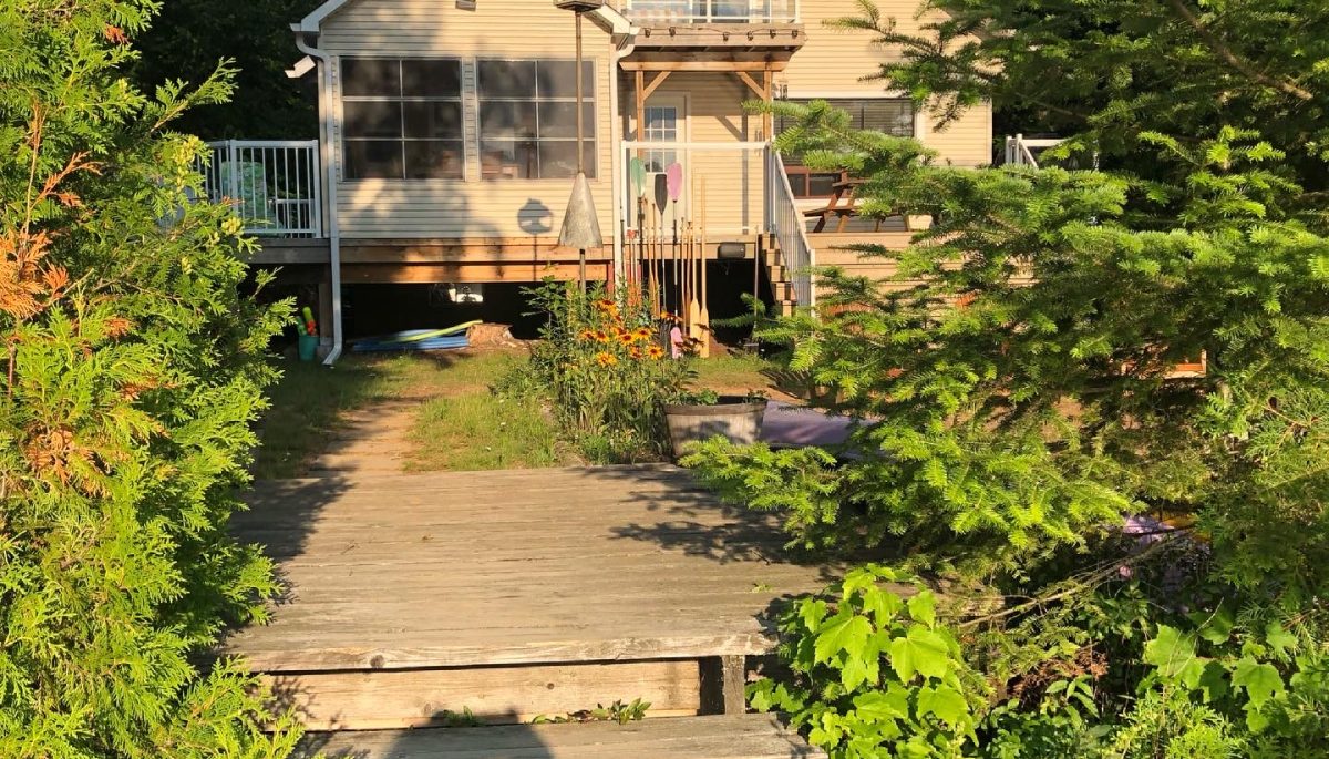 A two-story wood cottage set back among green trees with a wood deck