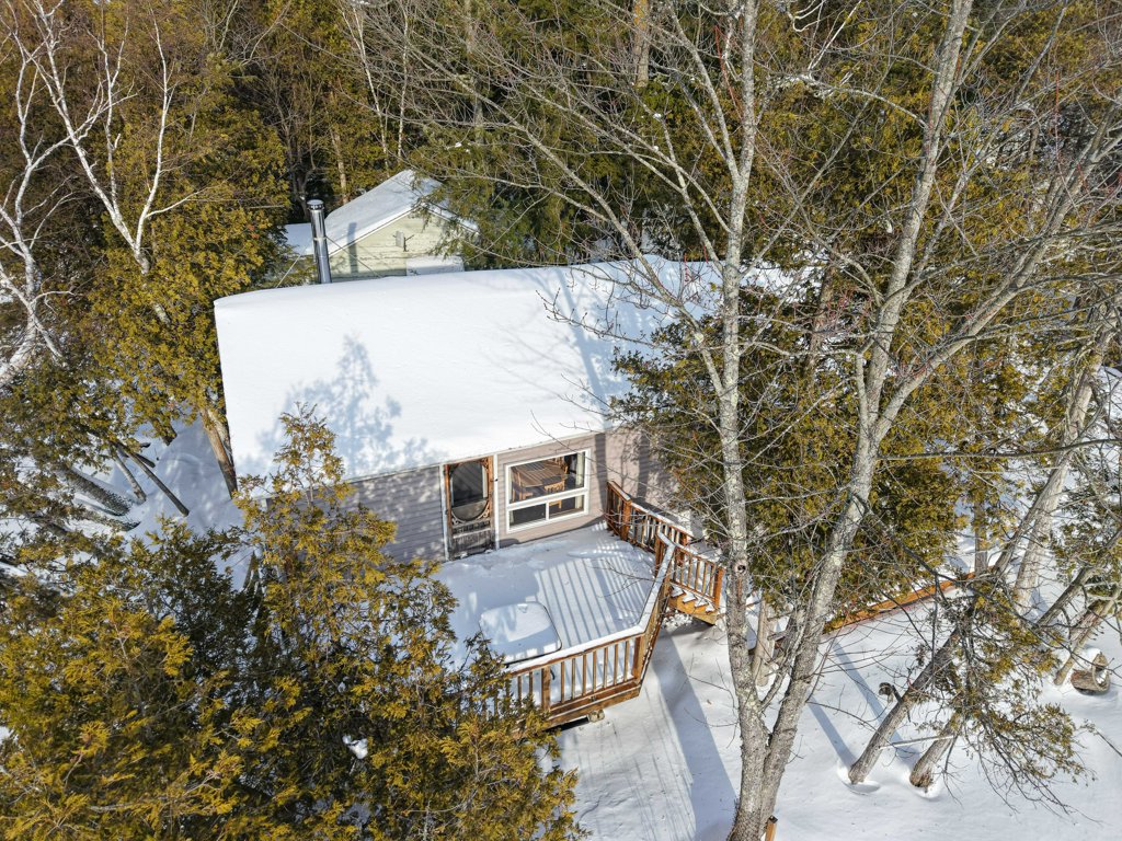 Drone view of one cottage with surrounding trees and an elevated deck near the waterfront.