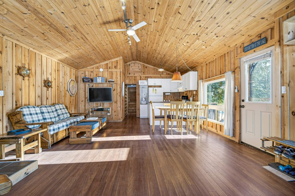 Bright open-concept cottage interior with wood-paneled walls, vaulted ceiling, living area, and dining space.
