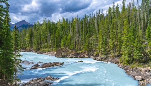 Turquoise water flows through the bow river natural bridge in banff national park, alberta, canada, surrounded by lush green pine trees and a cloudy sky