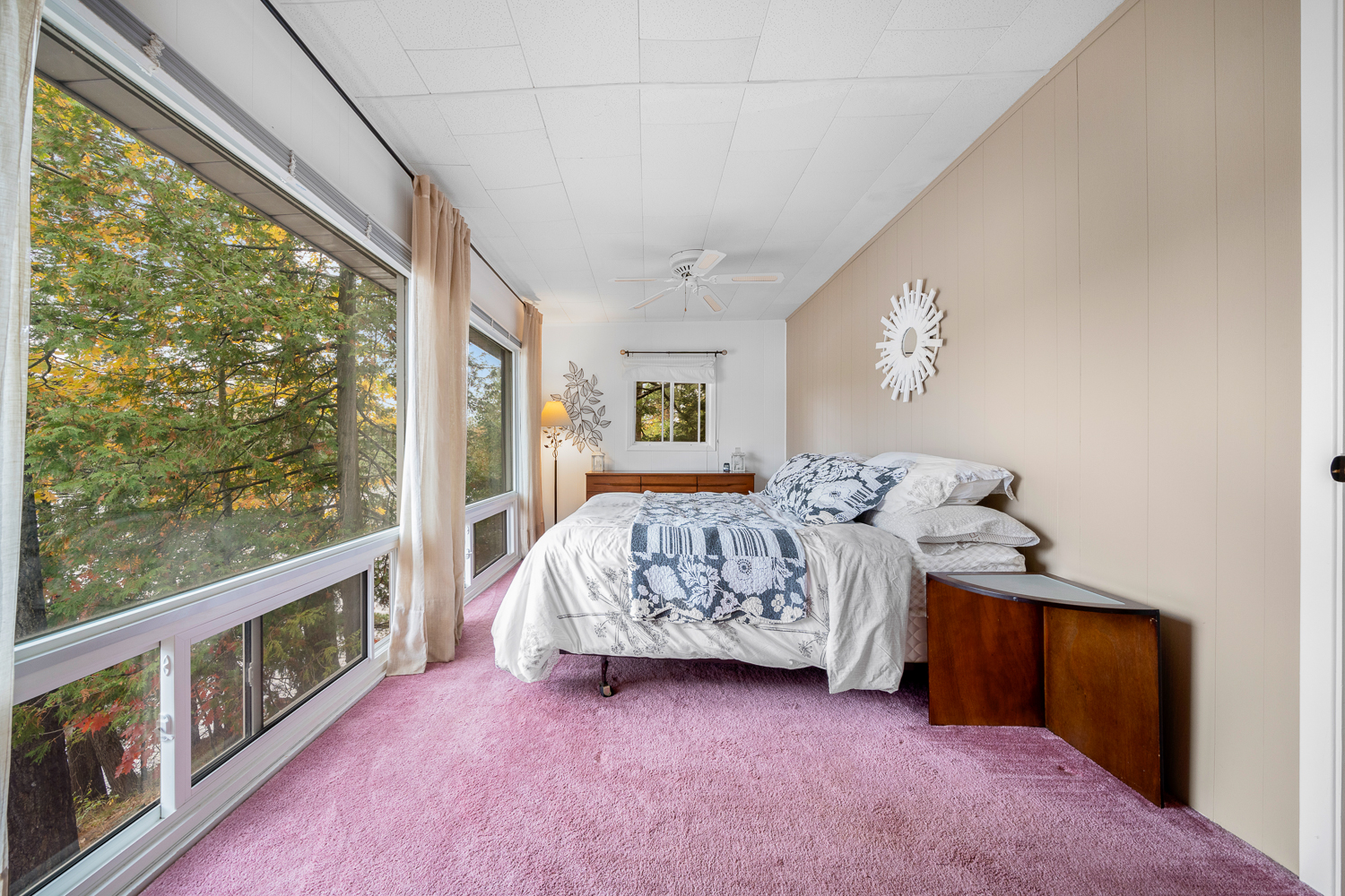 A white bed faces floor-to-ceiling windows. Pink carpeting covers the floor