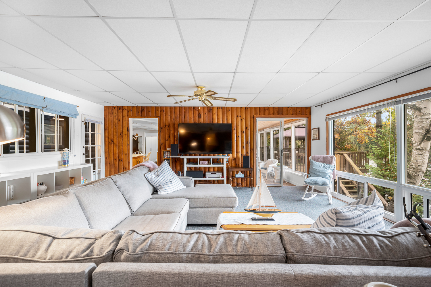 L-shaped beige couches face a wood-paneled wall with a TV in a sunny room