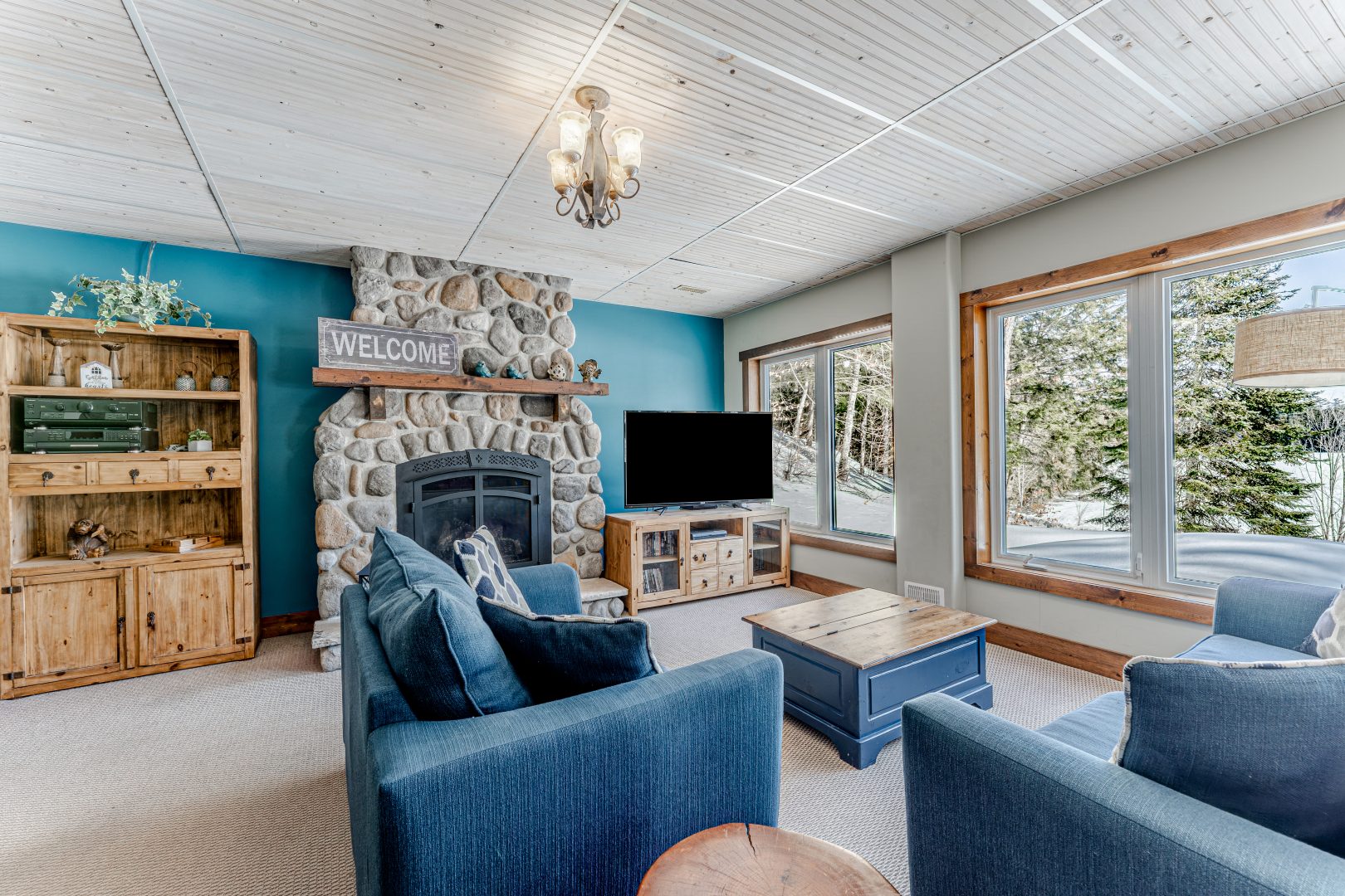 Blue couches face a TV and stone fireplace next to bright windows