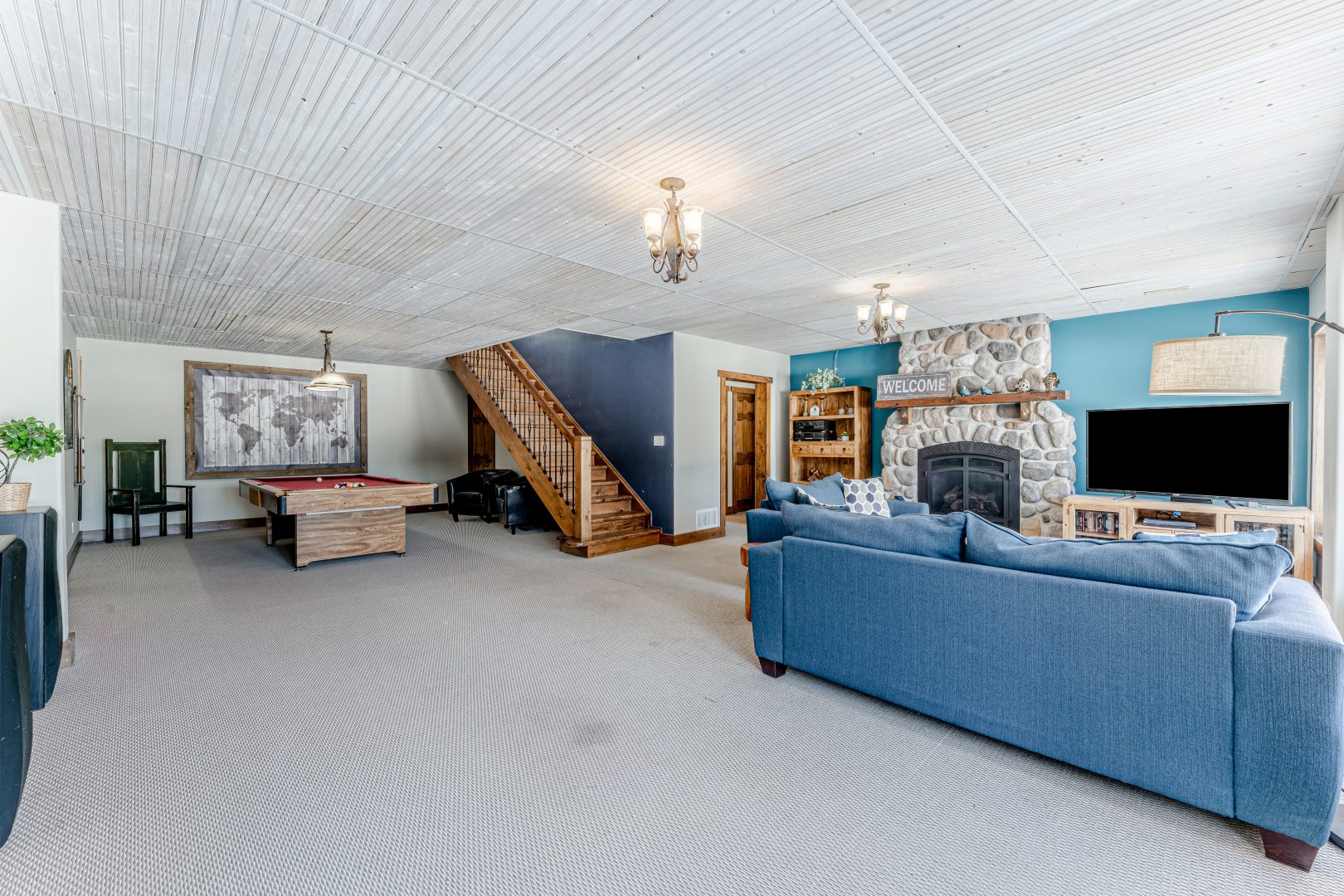 A pool table is tucked away beside wood stairs. To the right, a blue couch faces a stone fireplace