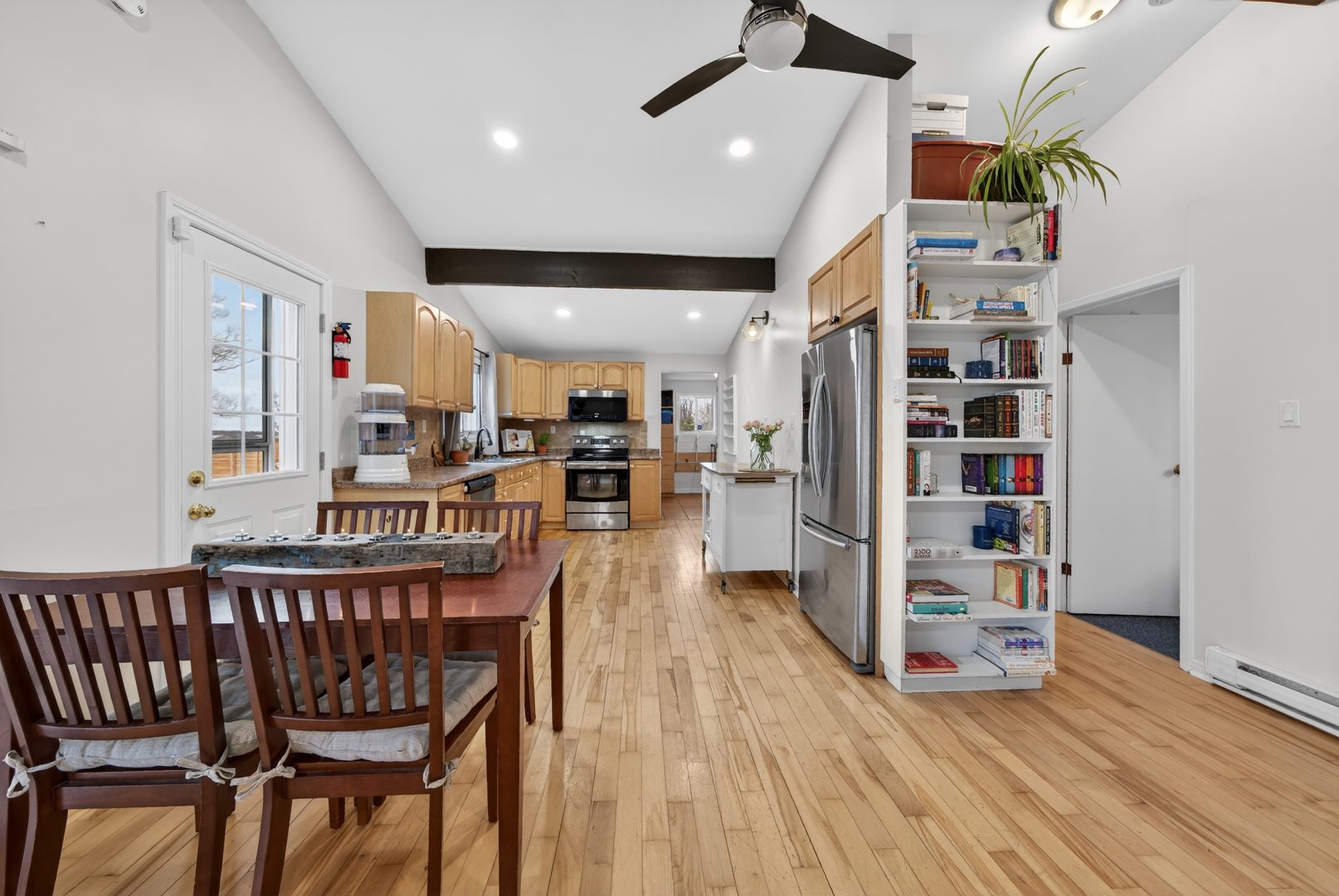 A white living area with light paneled flooring. A dark wood dining table is adjacent to a wood kitchen with stainless steel appliances