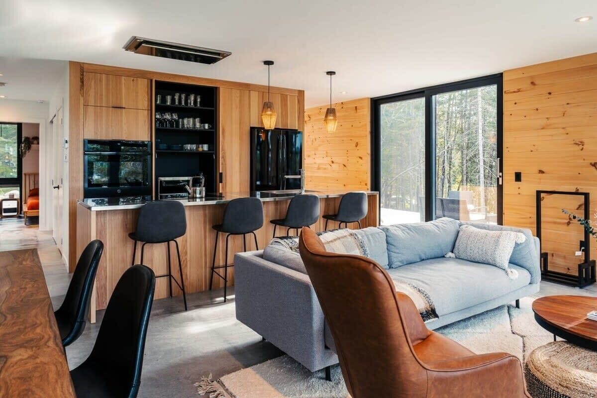 Kitchen and living room of the luxury chalet. Kitchen cabinets are wooden, with grey and brown couches in the living room and black stools across the kitchen island.
