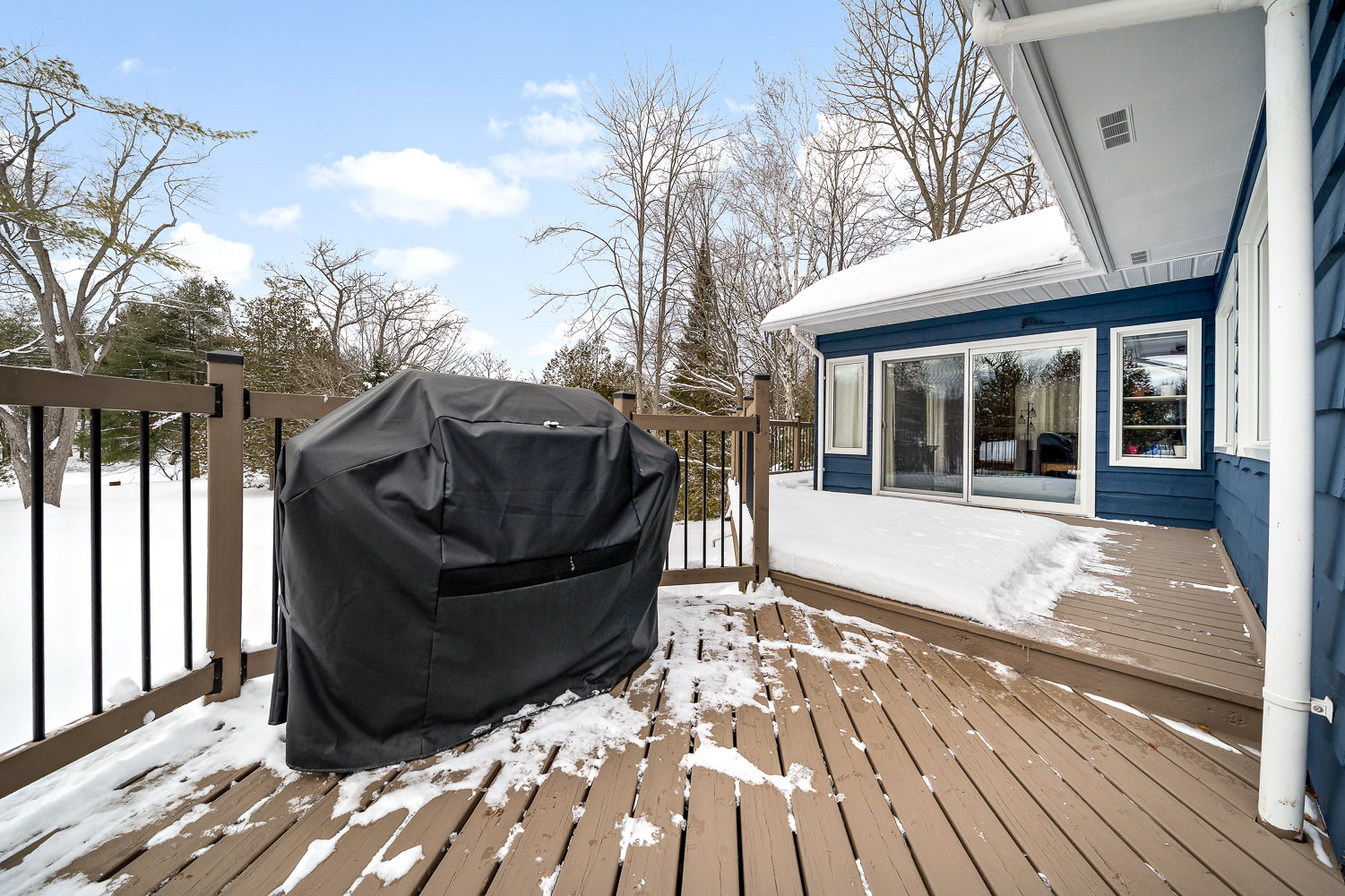A wood deck with a covered barbecue next to a snow-covered blue cottage