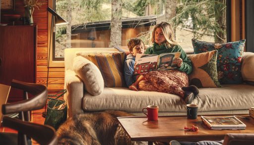 A mother reading a book to her son sitting on a beige couch with assorted coloured pillows
