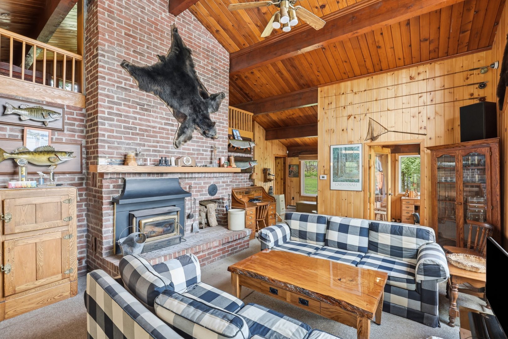 Blue and white plaid couches face each other. Beside them in a brick fireplace with a wood stove and a bear pelt hanging on the wall