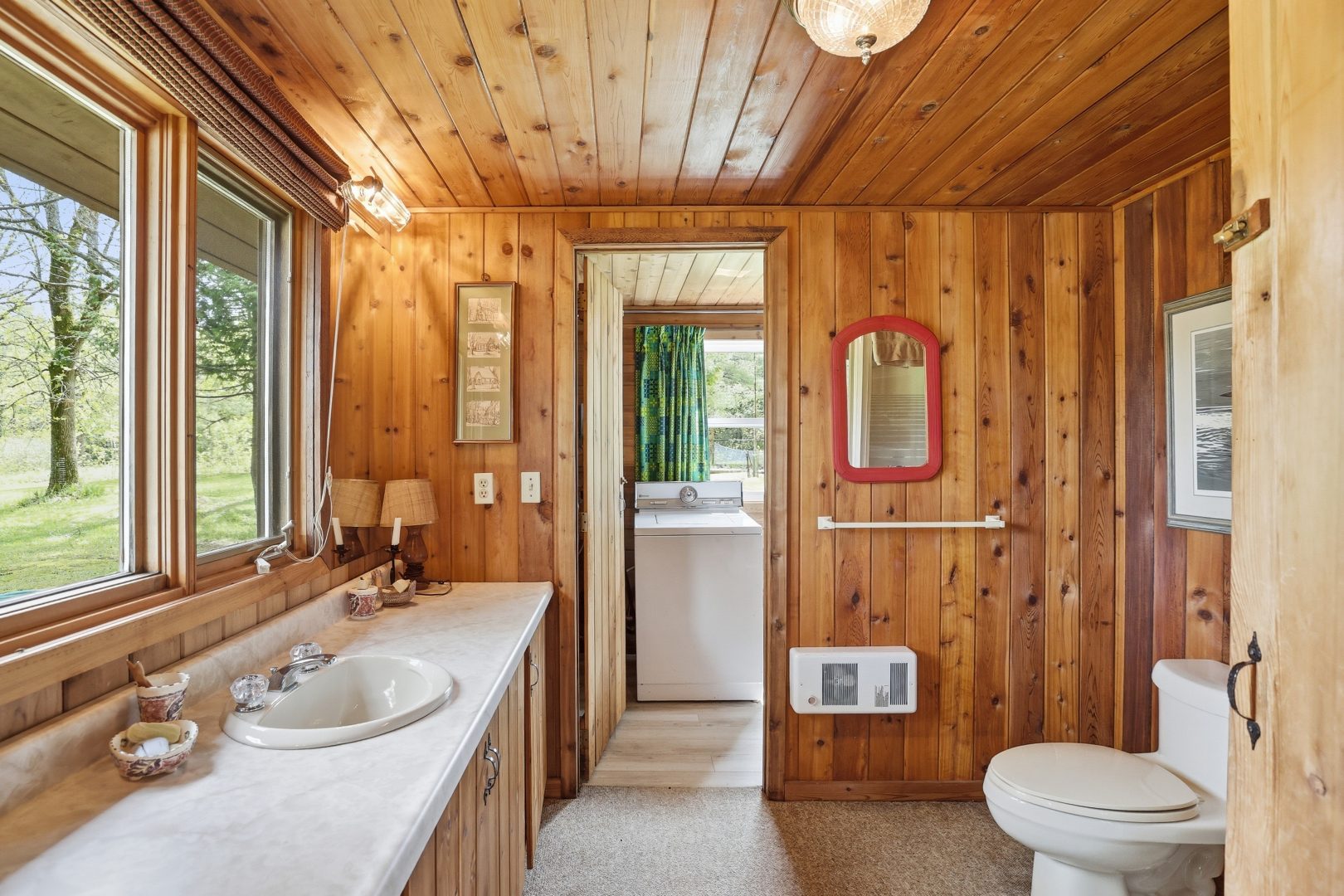 A wood panelled bathroom with a long white vanity. At the back, an open doorway reveals a white laundry machine
