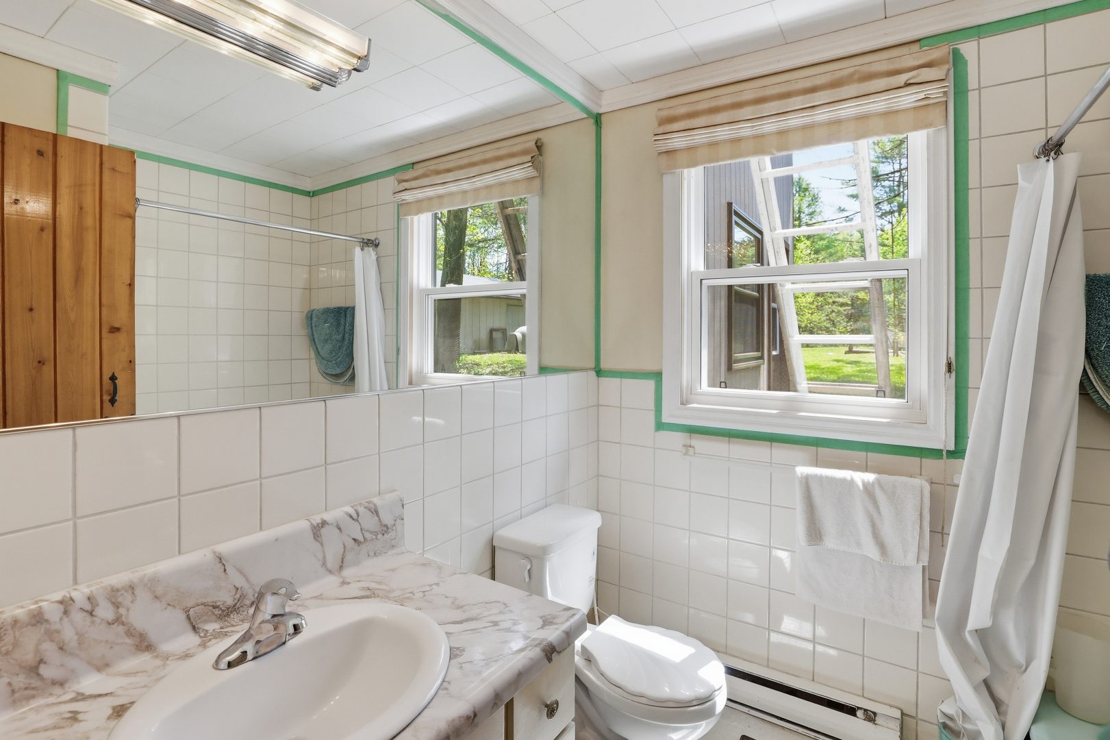 A white-tiled bathroom with a large mirror and a mirror looking out onto the grassy property