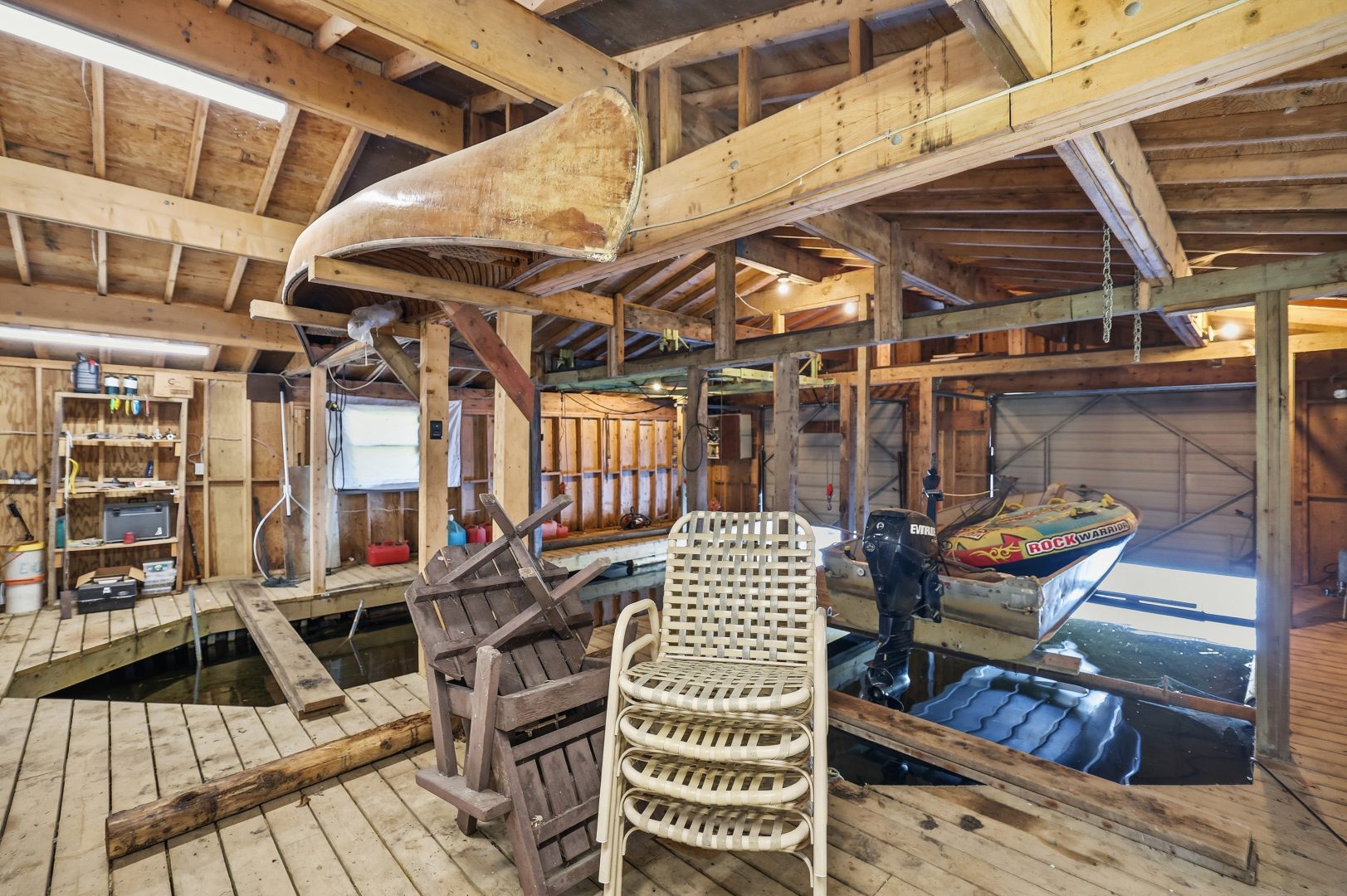 The inside of a boathouse with hanging canoes, stacked chairs, and a boat in the water
