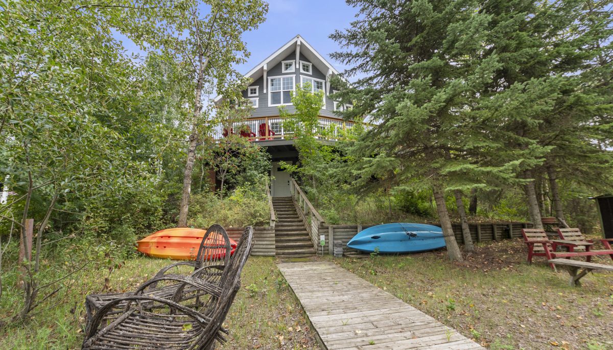 A grey cottage with a large balcony tucked away behind trees. A boardwalk leads down to a grassy area
