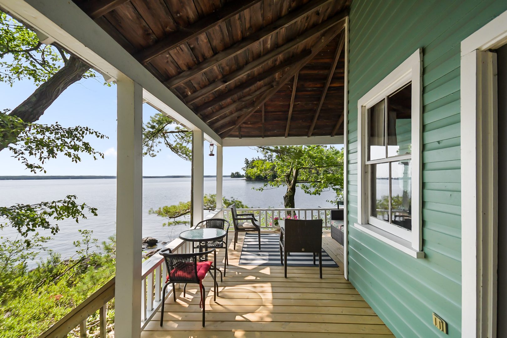 A covered porch along the side of a teal cottage facing the water