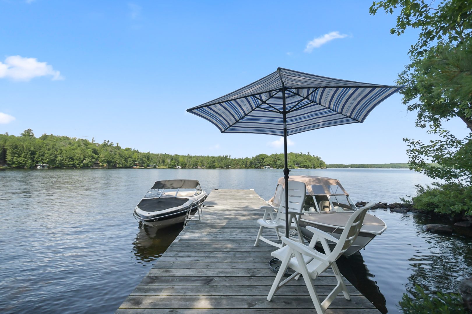 A dock with white chairs and a striped umbrella. A boat is parked at the end of the dock.