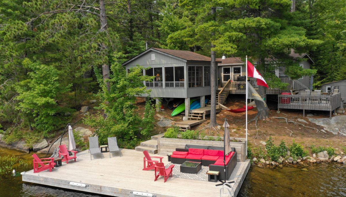 A grey cottage with multi-level decks on a forested property with a large dock