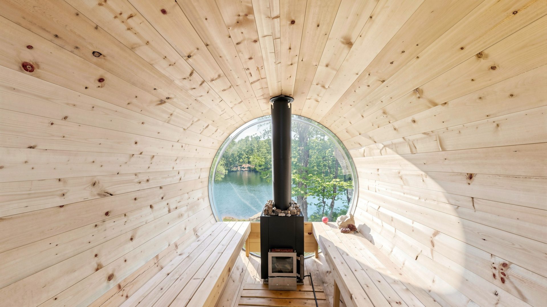 The interior of a light wood barrel sauna with a wood stove that looks out onto the lake