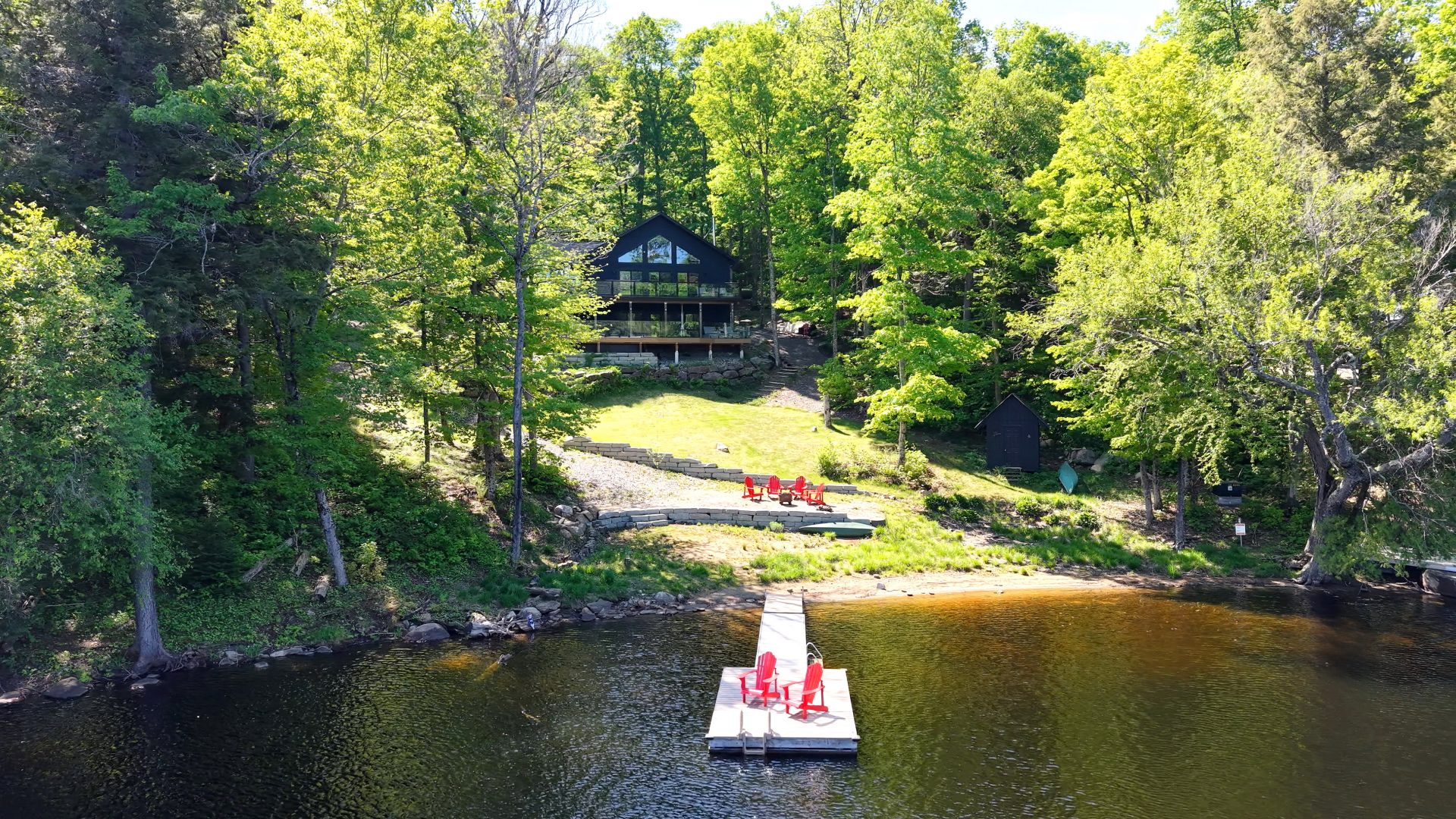 A black clapboard cottage is set back against a forest area with a sloping lawn. At the shoreline, a dock with red Muskoka chairs juts out into the water