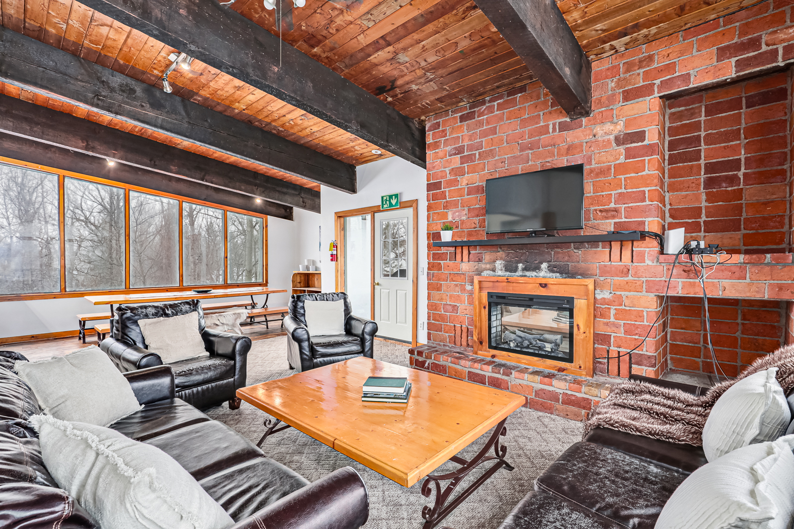 A spacious living room with black leather couches around a wood table. A fireplace set into a brick wall is on the adjoining wall