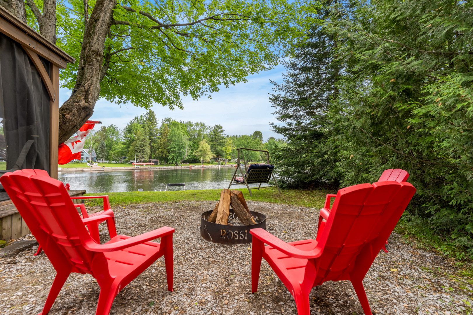 Red Muskoka chairs face a fire pit next to the water