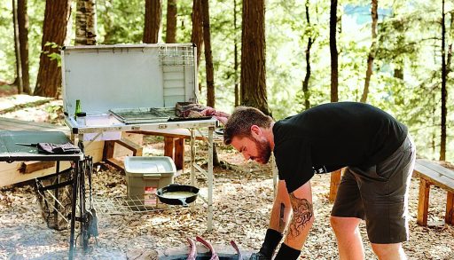 A man bent over a campfire cooking cuts of meat on a grill, sunlight through the trees in the background