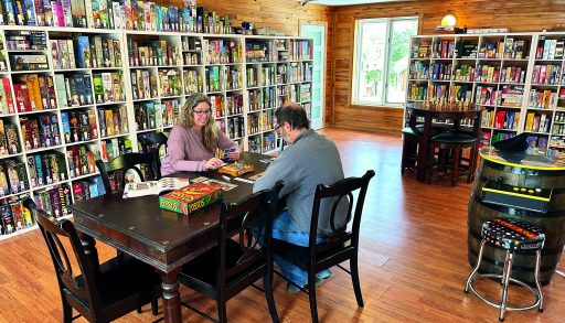 A couple play a board game on a table in a brightly lit room with many shelves of other board games