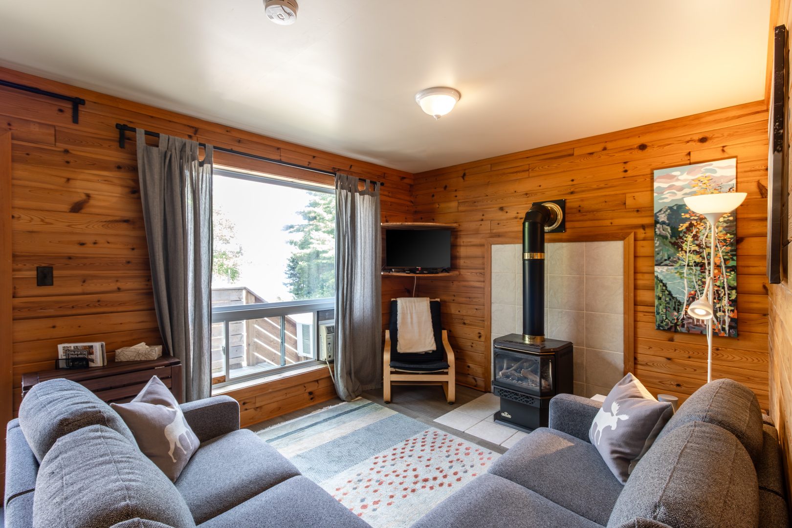 An L-shaped grey couch faces a wood stove in a wood-paneled room