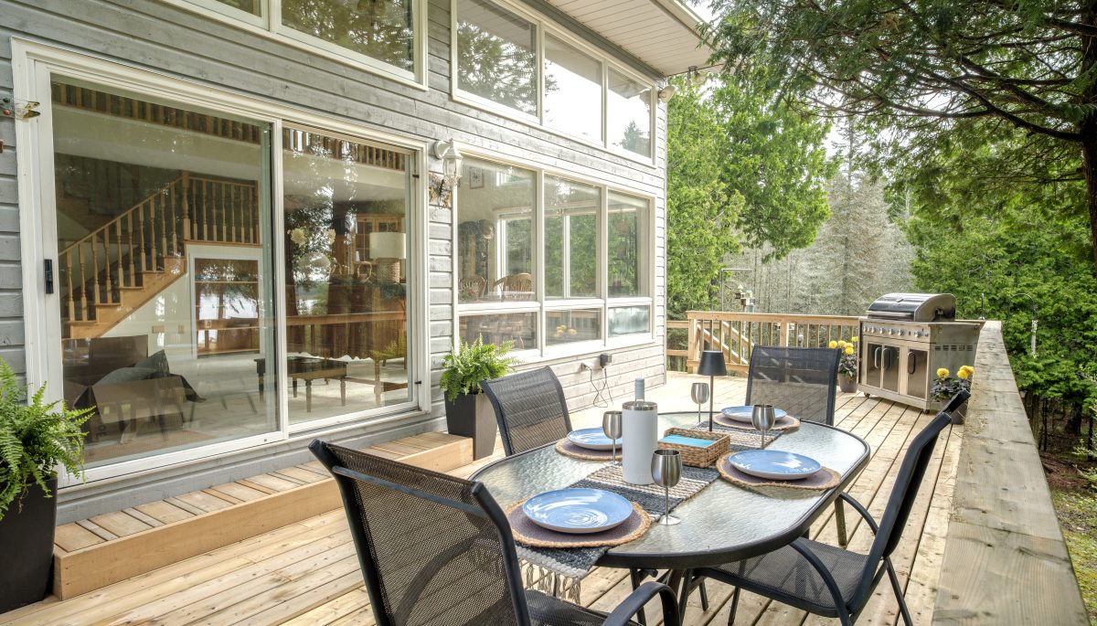 A patio dining set on a light wood deck outside a window-filled cottage