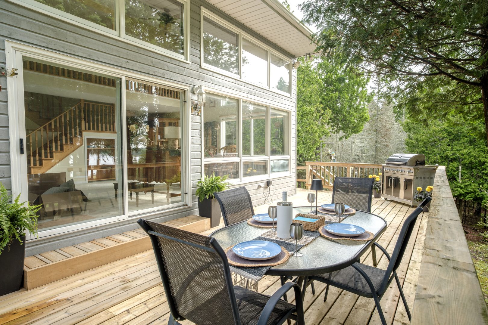 A patio dining set on a light wood deck outside a window-filled cottage