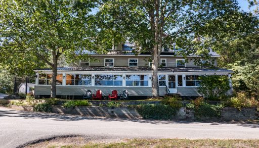 A long clapboard cottage with bright windows and a wraparound porch set back on a tree-lined property