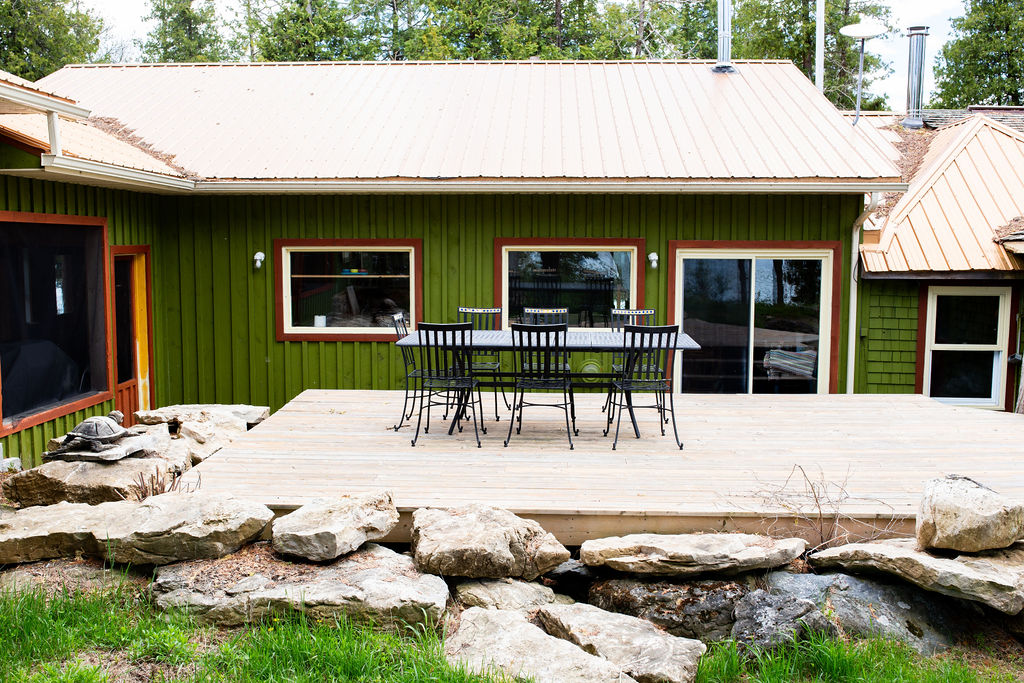 A black patio set on a light wood deck next to a green cottage