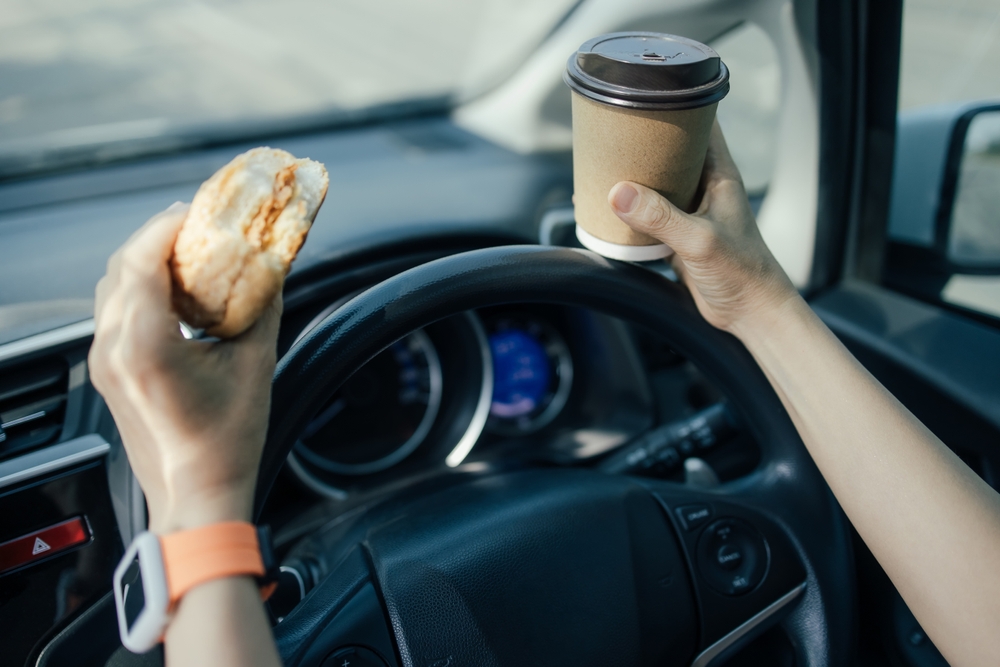 A woman driving while eating a sandwich and drinking coffee
