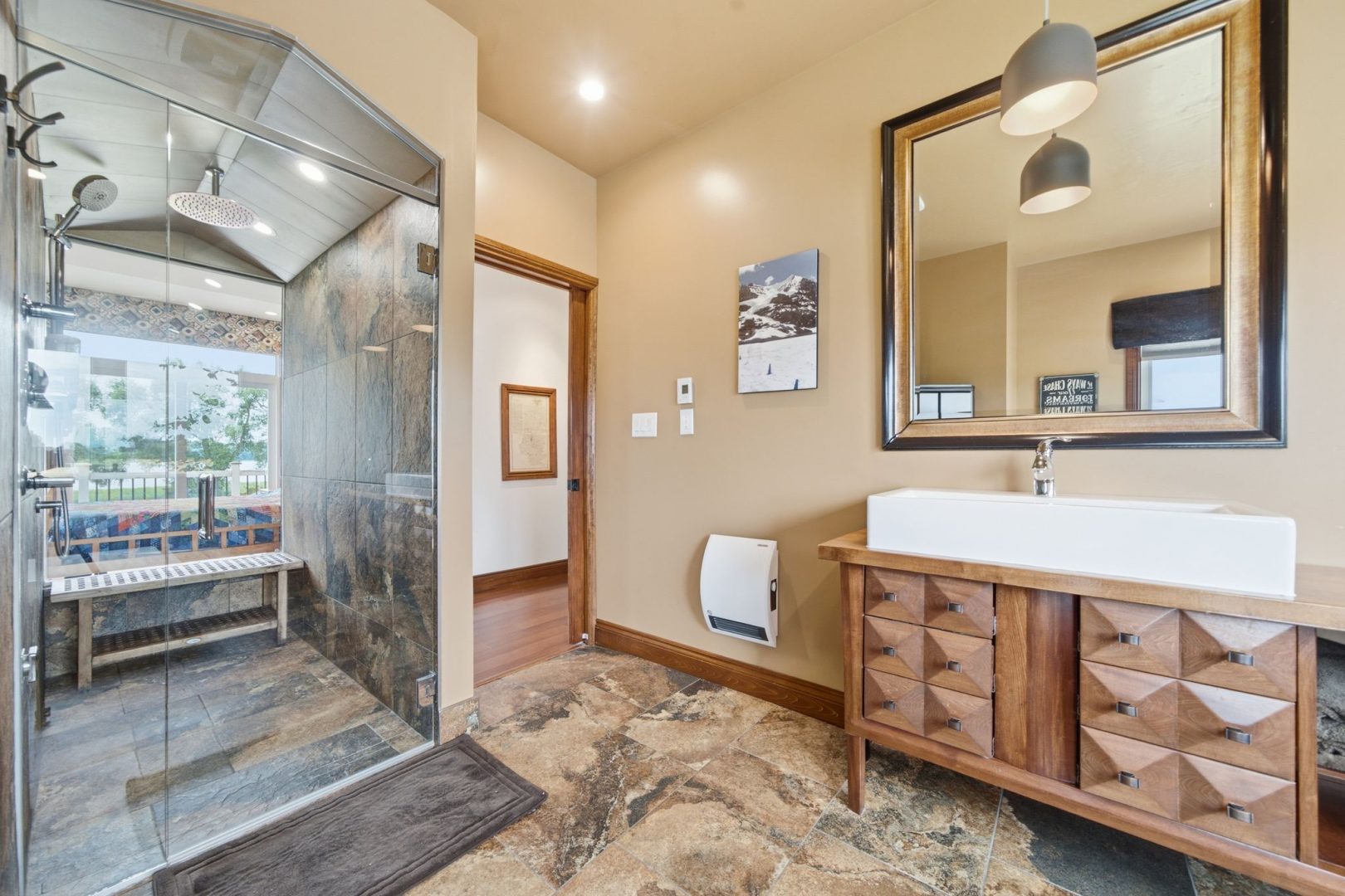 A beige bathroom with a glass walk-in shower, wood vanity and brown stone floors